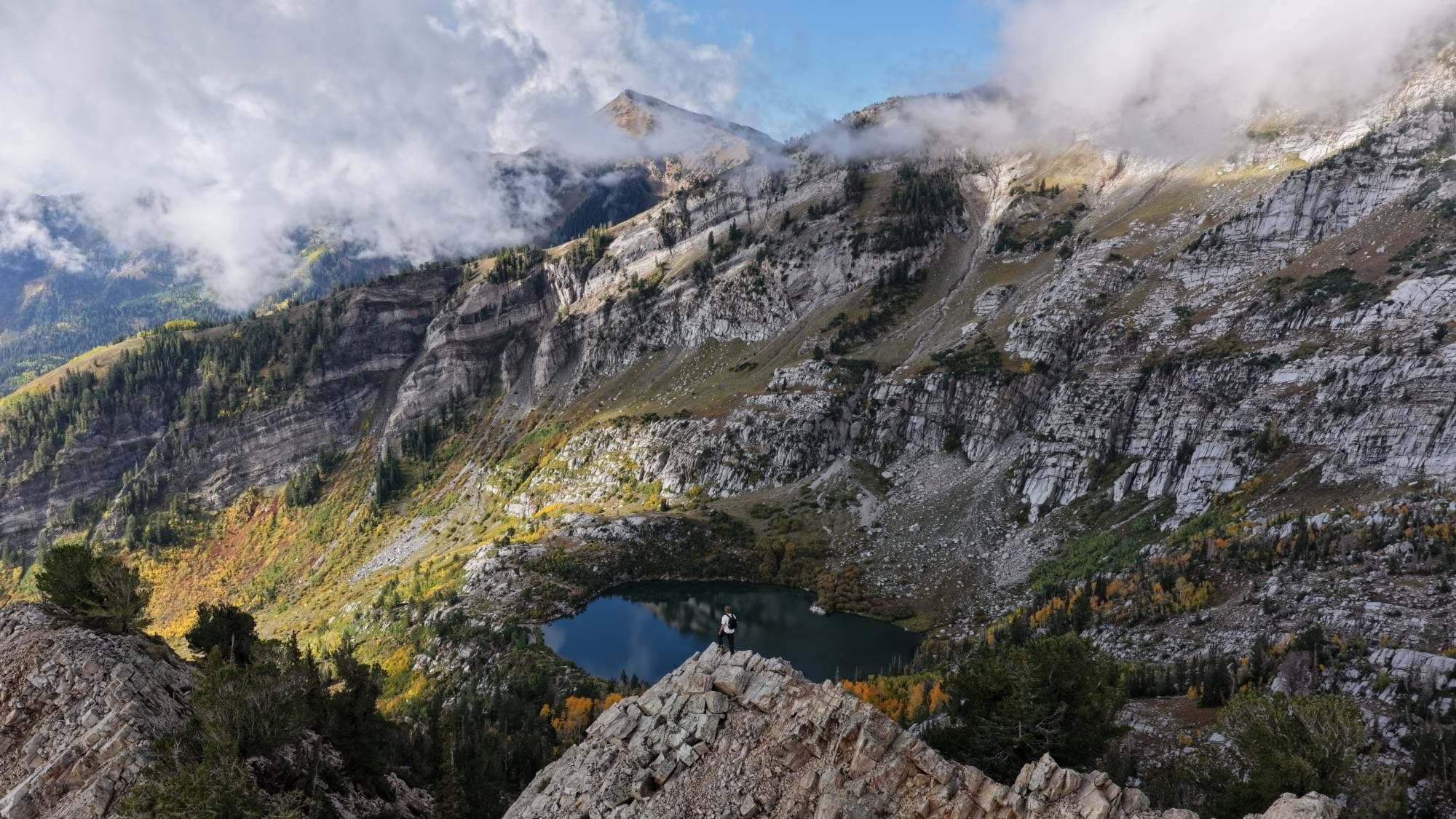 Russell standing on a rocky ridge above a mountain lake, with autumn color on the slopes and clouds drifting across the peaks