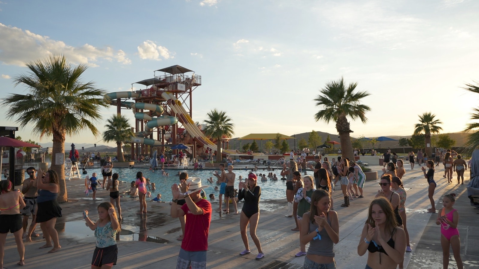 Guests joining an activity on the pool deck at golden hour, with the slide tower and palm trees behind them